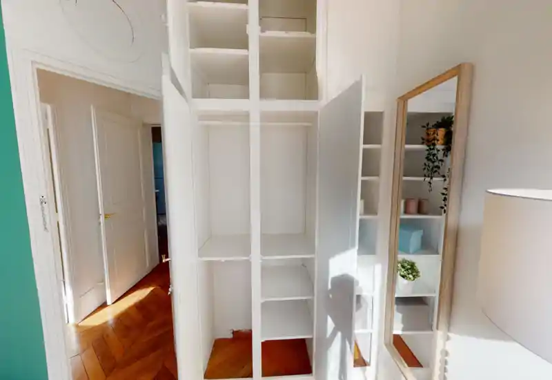 Bright bedroom area focused on a built-in white wardrobe with open doors and shelving; mirrored full-length mirror and wooden parquet floor add warmth and depth to the space.