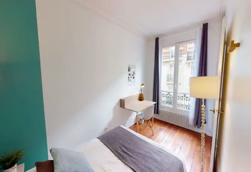 Bright, cozy bedroom with a comfortable double bed, a wall-mounted desk by a large window, and warm hardwood floors — ideal for remote work and restful nights.