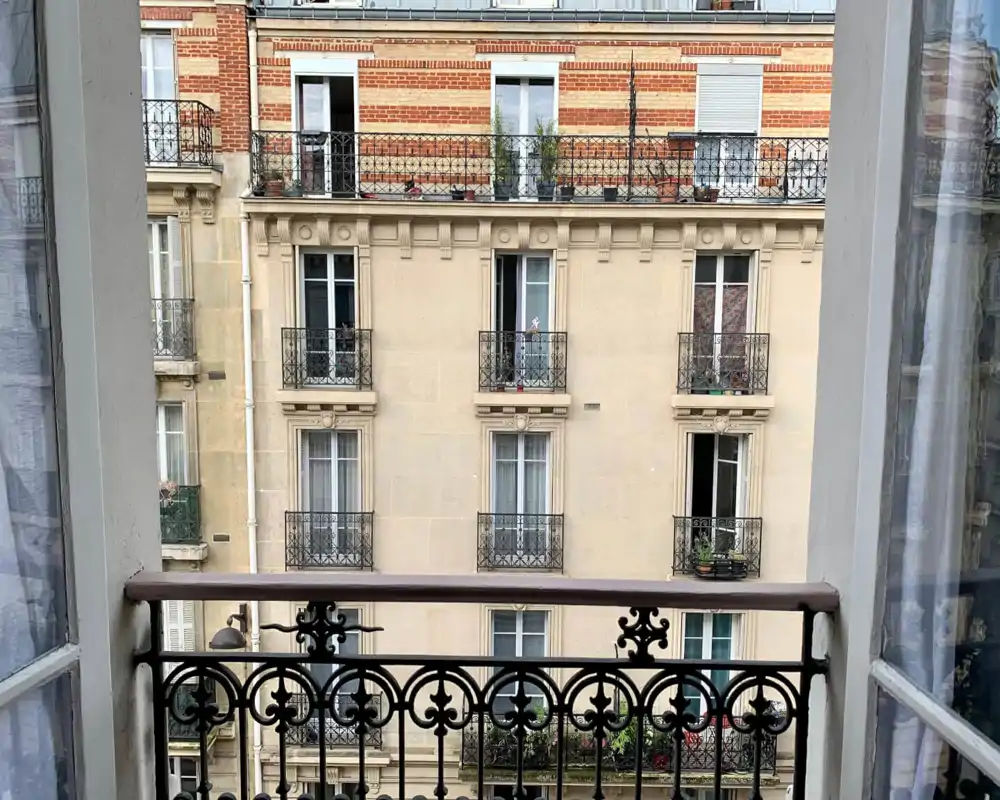 Vue depuis un petit balcon de style parisien sur un immeuble voisin avec garde-corps en fer forgé, pots de fleurs et façade en pierre classique.