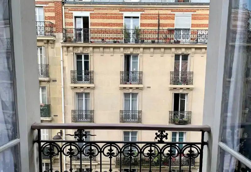 Vue depuis un petit balcon de style parisien sur un immeuble voisin avec garde-corps en fer forgé, pots de fleurs et façade en pierre classique.
