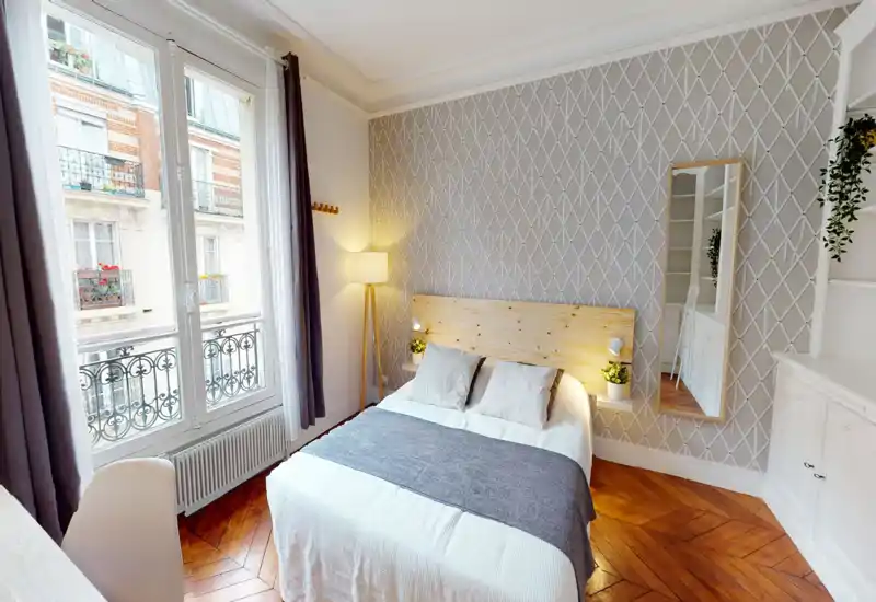 Chambre lumineuse et élégante avec tête de lit en bois, lit double en linge blanc et jeté gris, grande fenêtre avec balcon, mur décoratif et parquet chaleureux.