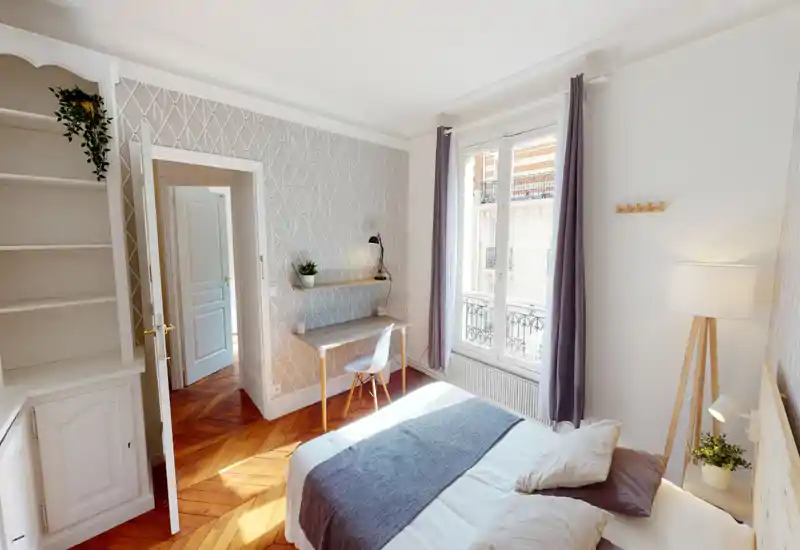 Chambre lumineuse et soignée avec un lit double, parquet chaleureux, petit bureau près de la fenêtre et belle lumière naturelle venant d’une grande fenêtre française.