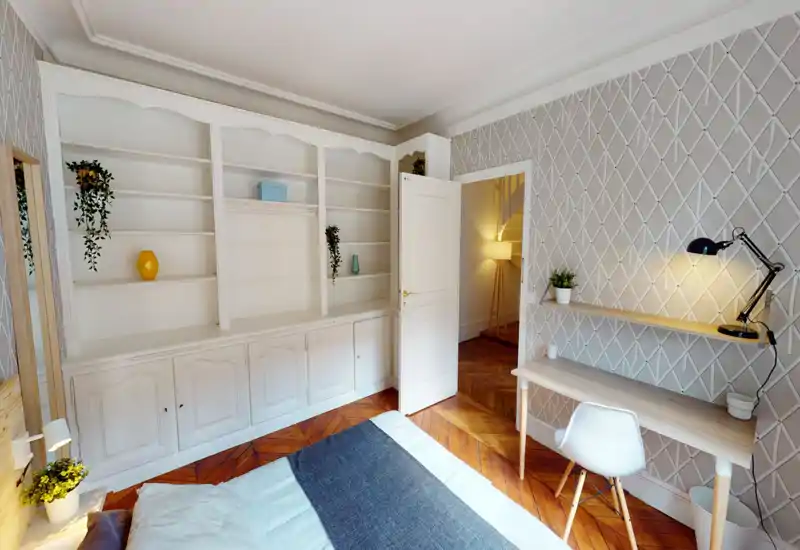 Chambre lumineuse et élégante avec lit bien visible au premier plan, étagères blanches intégrées, parquet et un petit bureau avec lampe de lecture—accueillante et soignée pour une annonce.