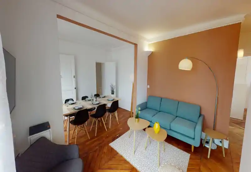 Salon lumineux et élégant avec un canapé trois places bleu-vert, tables basses gigognes, tapis à motifs et lampadaire arche ; on aperçoit la salle à manger ouverte à l'arrière, offrant un espace convivial et chaleureux.
