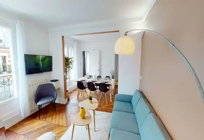 Séjour lumineux et élégant avec canapé bleu-vert, lampadaire moderne et petites tables basses; coin repas visible à travers une ouverture, grandes fenêtres apportant beaucoup de lumière naturelle.
