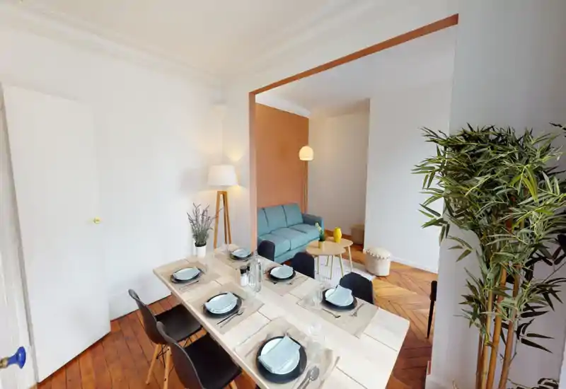 Salle à manger lumineuse et décorée avec une grande table en bois dressée pour six, chaises noires modernes et vue sur un coin salon cosy avec canapé turquoise et mur d'accent chaleureux.