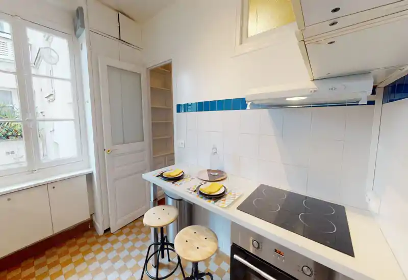 Cuisine compacte et lumineuse avec plaque de cuisson, four encastré et un petit bar pour le petit-déjeuner avec deux tabourets en bois. La grande fenêtre apporte une belle lumière naturelle; crédence carrelée et sol à motifs donnent un charme vintage.