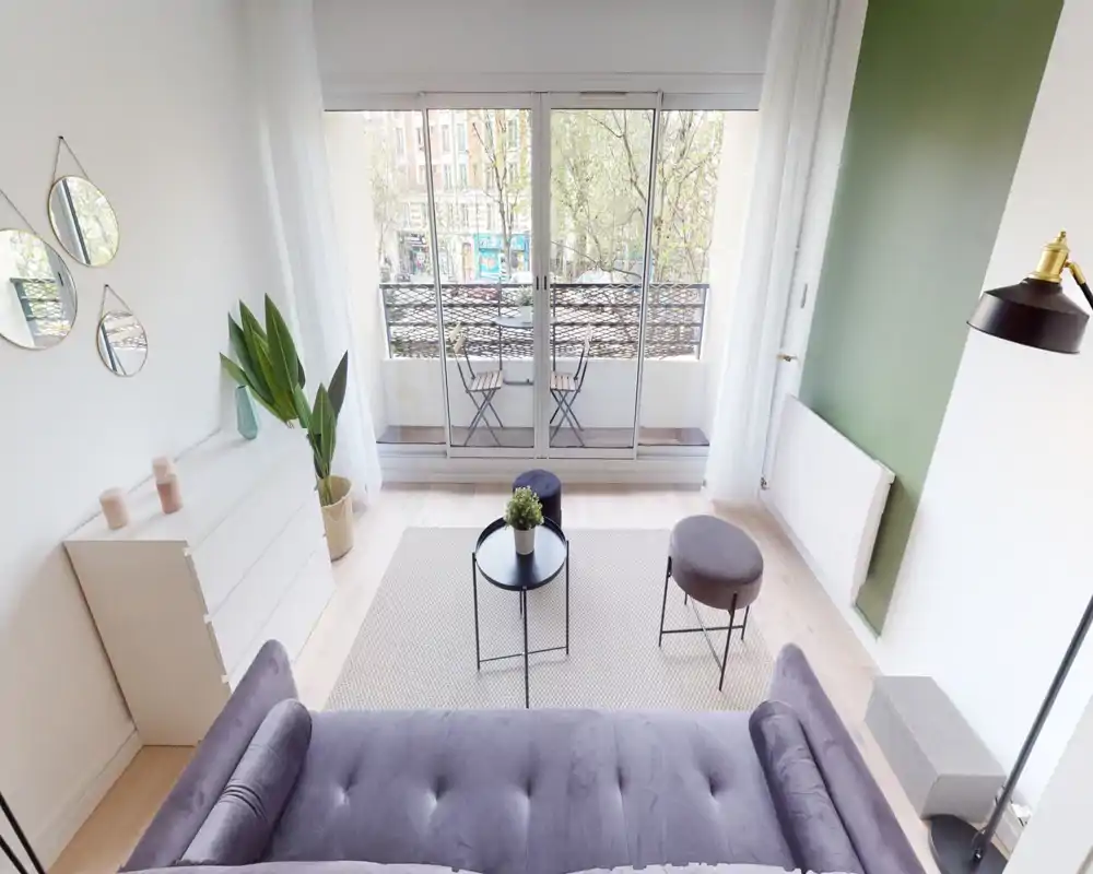 Salon moderne et lumineux avec canapé violet, table basse, poufs et grandes baies vitrées donnant sur un petit balcon. Parquet clair, tapis neutre et mur vert apportent une ambiance apaisante et stylée.