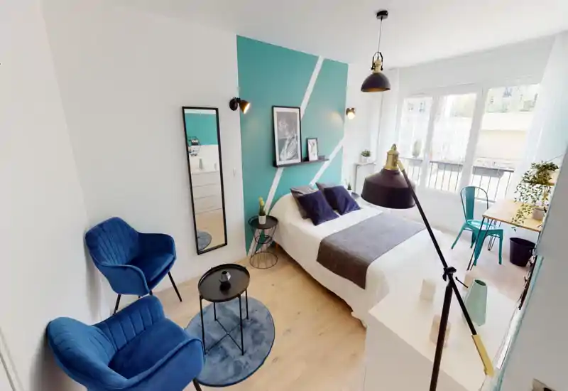 Bright, stylish bedroom with a double bed, decorative pillows and a teal feature wall; large window provides abundant natural light and a small desk by the window creates a multifunctional space.