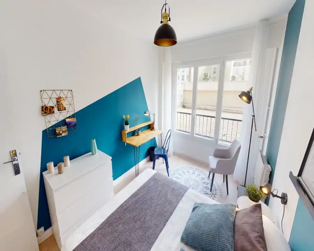 Chambre lumineuse et élégante avec un mur d'accent bleu-vert, grande fenêtre et lit bien fait ; comprend un petit bureau, un fauteuil confortable et un éclairage moderne.