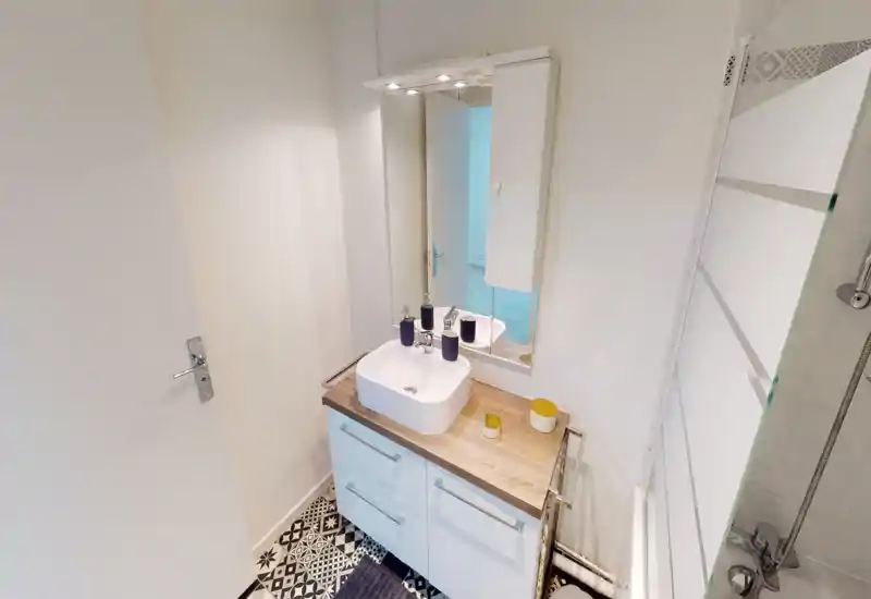 Compact modern bathroom with a countertop sink, wall mirror and a shower area with glass screen; patterned floor tiles add visual character.