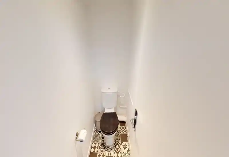 Compact, clean WC with patterned tile floor and dark wooden toilet seat set against white walls — well-lit and tidy.