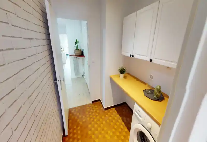 Compact utility/laundry area with a wooden countertop, white overhead cabinets and a washing machine; bright, clean space with small decorative plants.
