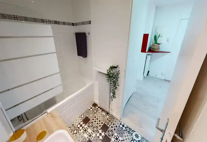 Bright, modern bathroom with patterned floor tiles, a built-in bathtub with half-glass screen, white tiled walls and a small radiator with a trailing plant.