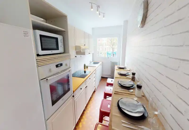 Bright, narrow kitchen with built-in oven, microwave, electric cooktop and a long breakfast bar set for meals. White cabinets and a window at the end make the space feel airy while red stools add a pop of color.