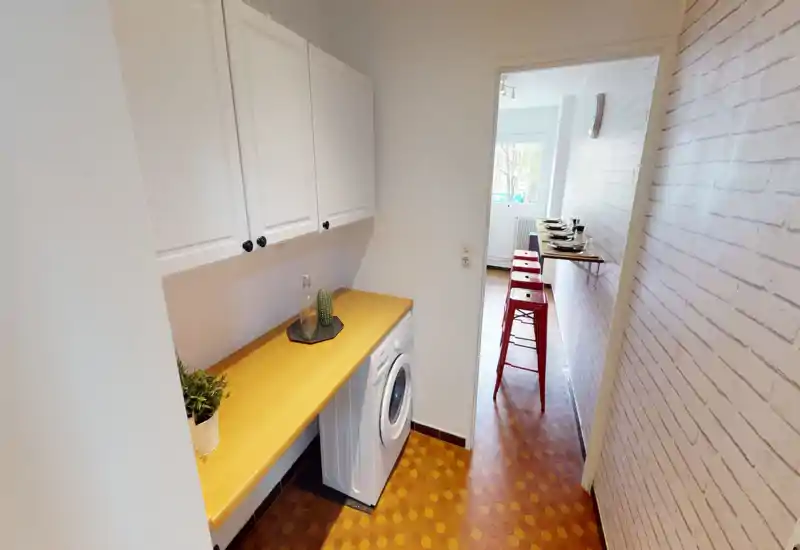 Compact kitchenette / laundry alcove with built-in countertop, upper cabinets and a front-loading washer. The space opens onto a dining bar with red stools and benefits from bright natural light.