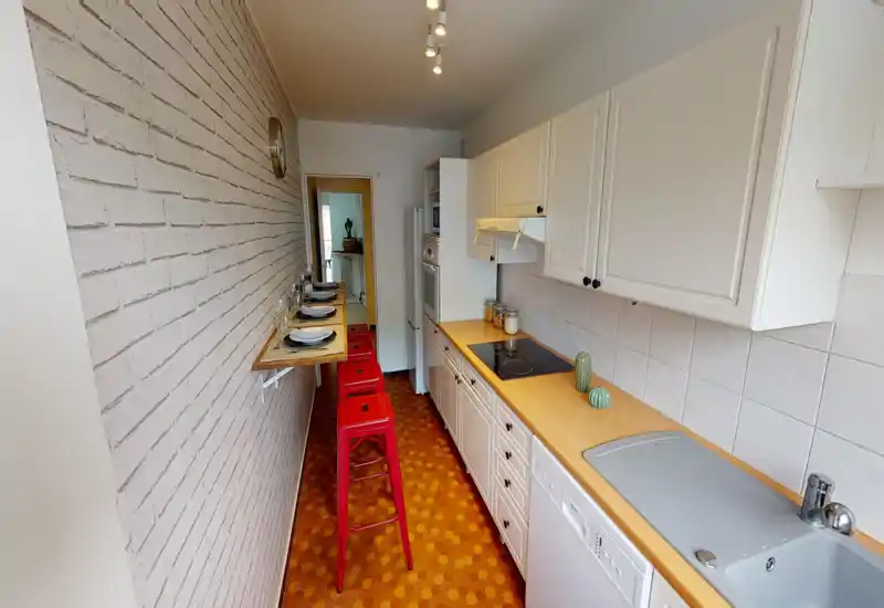 Narrow galley kitchen with white cabinets, wooden countertops, electric cooktop and integrated appliances. A fold-down breakfast bar with red stools and simple table settings gives a cozy, practical feel.