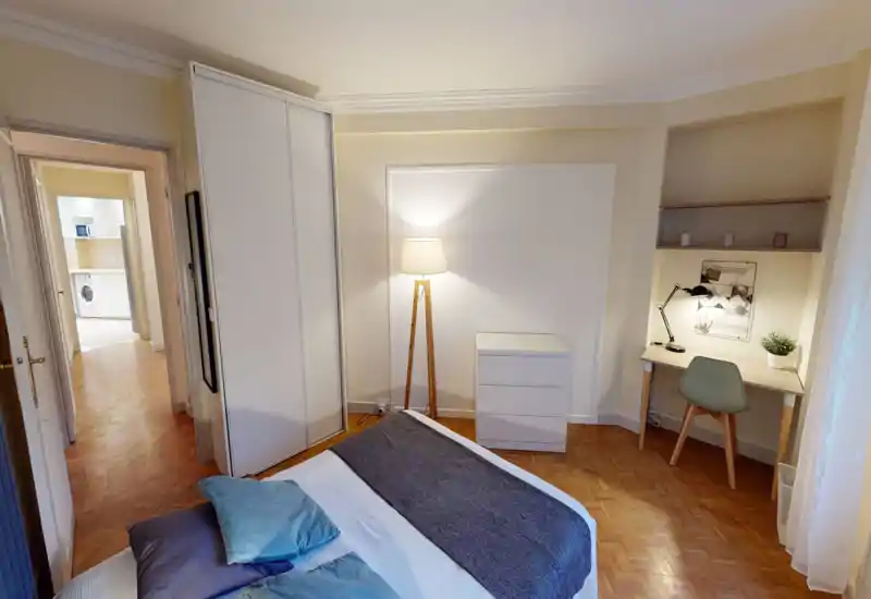 Chambre bien mise en scène avec lit double, lampe, armoire et un coin bureau confortable ; tons chauds et parquet pour une ambiance accueillante.