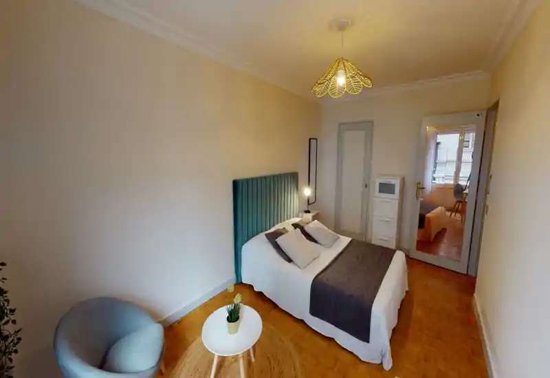 Bright, cozy bedroom with a teal upholstered headboard, neatly made double bed, bedside lamp and a comfortable armchair by a small round table.