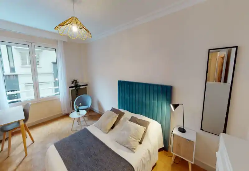 Bright, stylish bedroom with a teal upholstered headboard, double bed dressed in neutral linens, bedside lamp and a full-length mirror. Large window provides abundant natural light and a cozy seating corner with a small table.