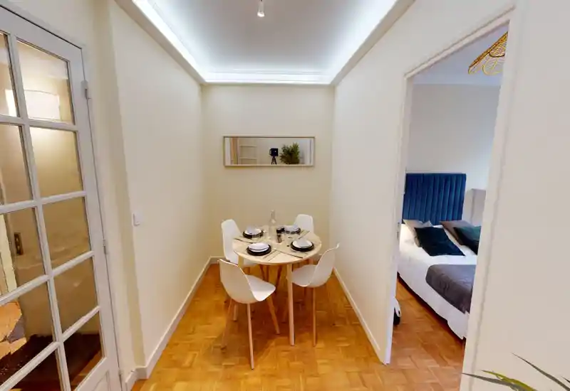 Compact dining area with a round wooden table set for four, modern white chairs and warm parquet flooring; bright, cozy space ideal for small meals.