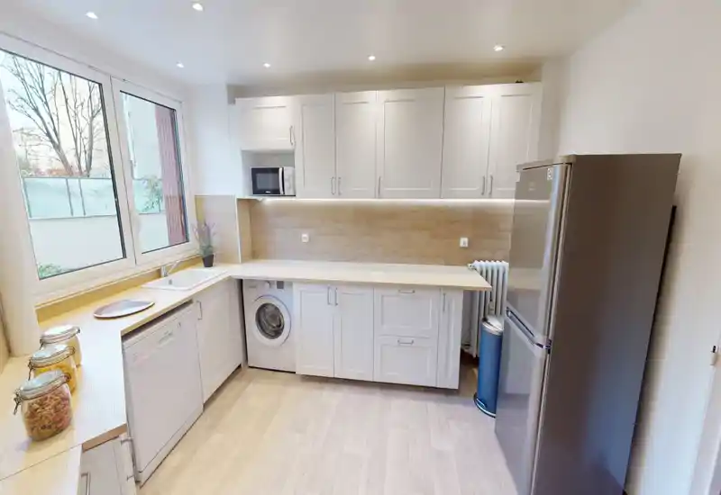 Bright modern kitchen with white cabinetry, a large window providing natural light, integrated appliances (dishwasher, washing machine, microwave) and a stainless-steel fridge — a clean, welcoming space for cooking and everyday use.
