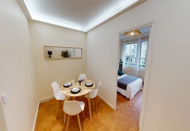 Compact dining nook with a round table set for four, modern white chairs and warm parquet flooring. The space opens onto a bedroom through a doorway and benefits from soft ceiling lighting and natural light from the adjacent room.