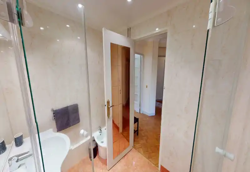 Bright, tiled bathroom with sink, bidet and a glass shower door; warm marble-look tiles and mirrored door add depth.