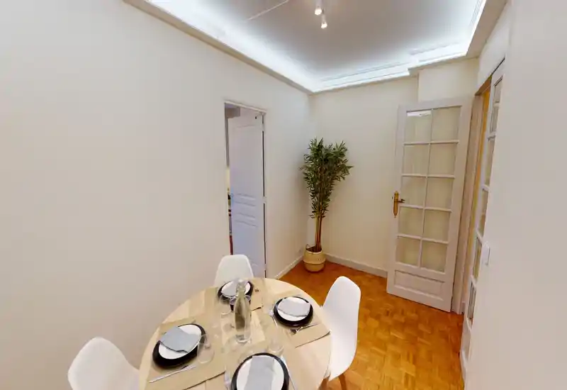 Bright, compact dining area with a round wooden table set for four and modern white chairs, warm parquet flooring and subtle ceiling lighting.