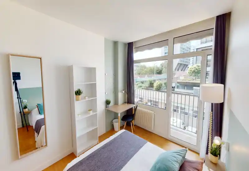 Chambre lumineuse et bien décorée avec une grande fenêtre et un lit confortable agrémenté de coussins et d'un plaid; un petit bureau près de la fenêtre et des plantes apportent une atmosphère chaleureuse et fonctionnelle.