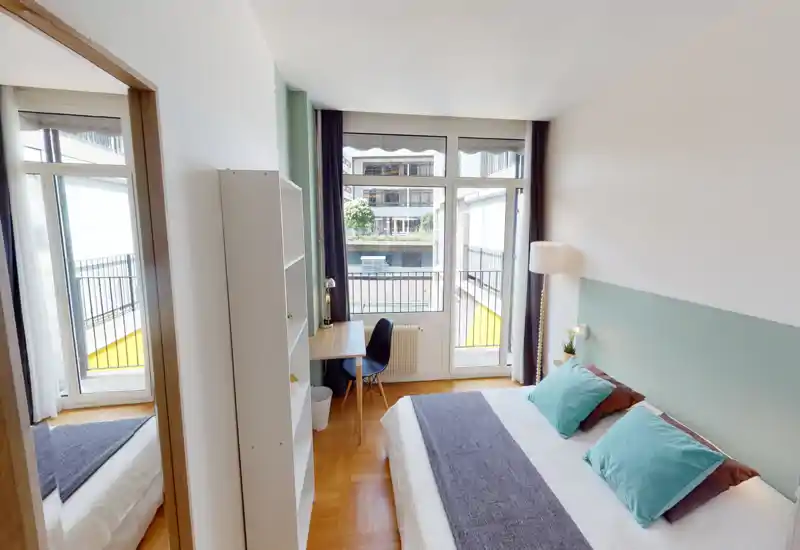 Chambre moderne et lumineuse avec grand lit double, lampe de chevet et coussins, bureau près de la fenêtre et accès direct à un petit balcon. Grandes fenêtres apportant une belle lumière naturelle et vue urbaine.