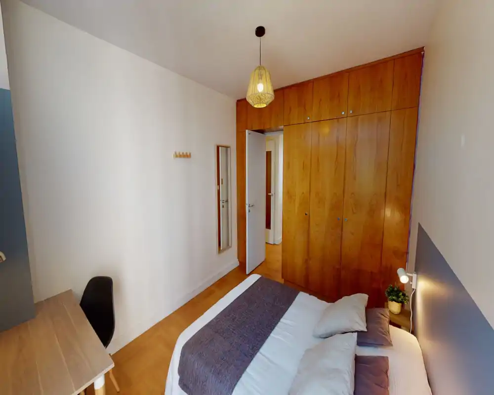 Chambre propre et lumineuse avec lit double bien habillé, lampe de chevet et grand placard en bois intégré. Les tons chauds et la lumière naturelle offrent un espace de nuit confortable, parfait pour une annonce locative.