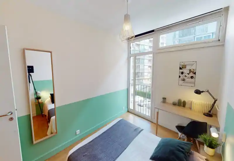 Chambre lumineuse et soignée avec un lit au premier plan, un petit bureau en bois près de la fenêtre et des accents contemporains comme des plantes et un miroir sur pied.