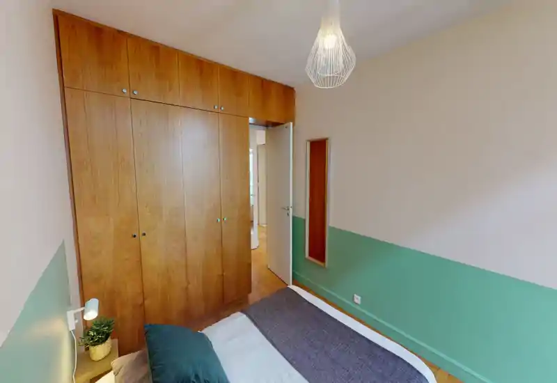 Chambre lumineuse et chaleureuse avec une grande armoire encastrée en bois, mur bas vert-bleu et un lit bien fait avec coussins — idéale pour un séjour confortable.