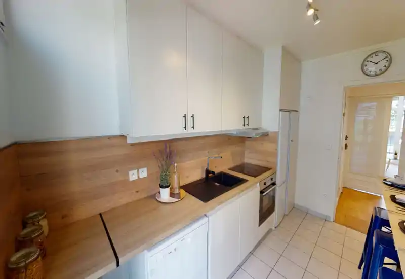 Cuisine moderne et compacte avec plan de travail et crédence imitation bois, four encastré, plaque à induction et évier intégré. Espace lumineux et propre avec des meubles blancs et de petites touches décoratives comme une plante en pot et des bocaux.