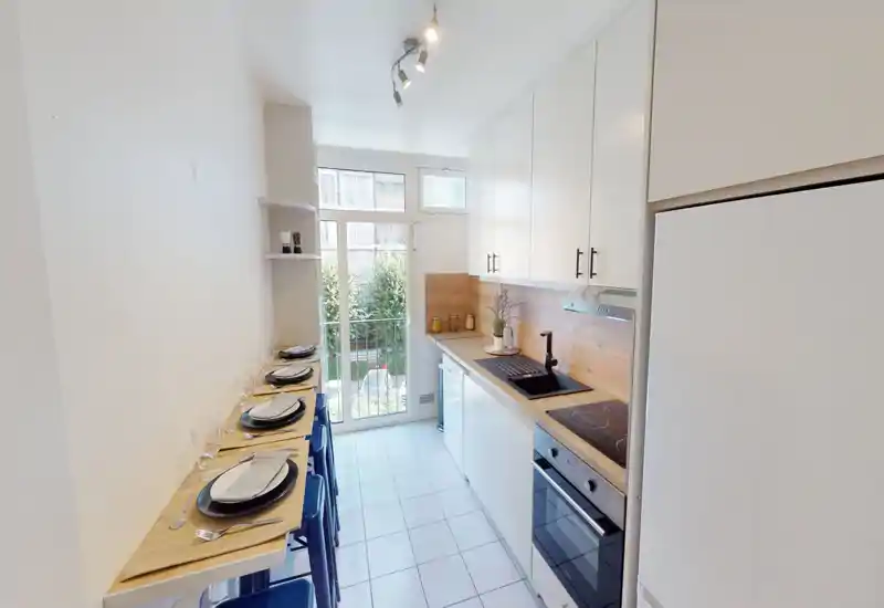 Cuisine compacte et lumineuse avec placards blancs, plan de travail en bois, four encastré et un bar étroit dressé pour quatre face à une porte vitrée.