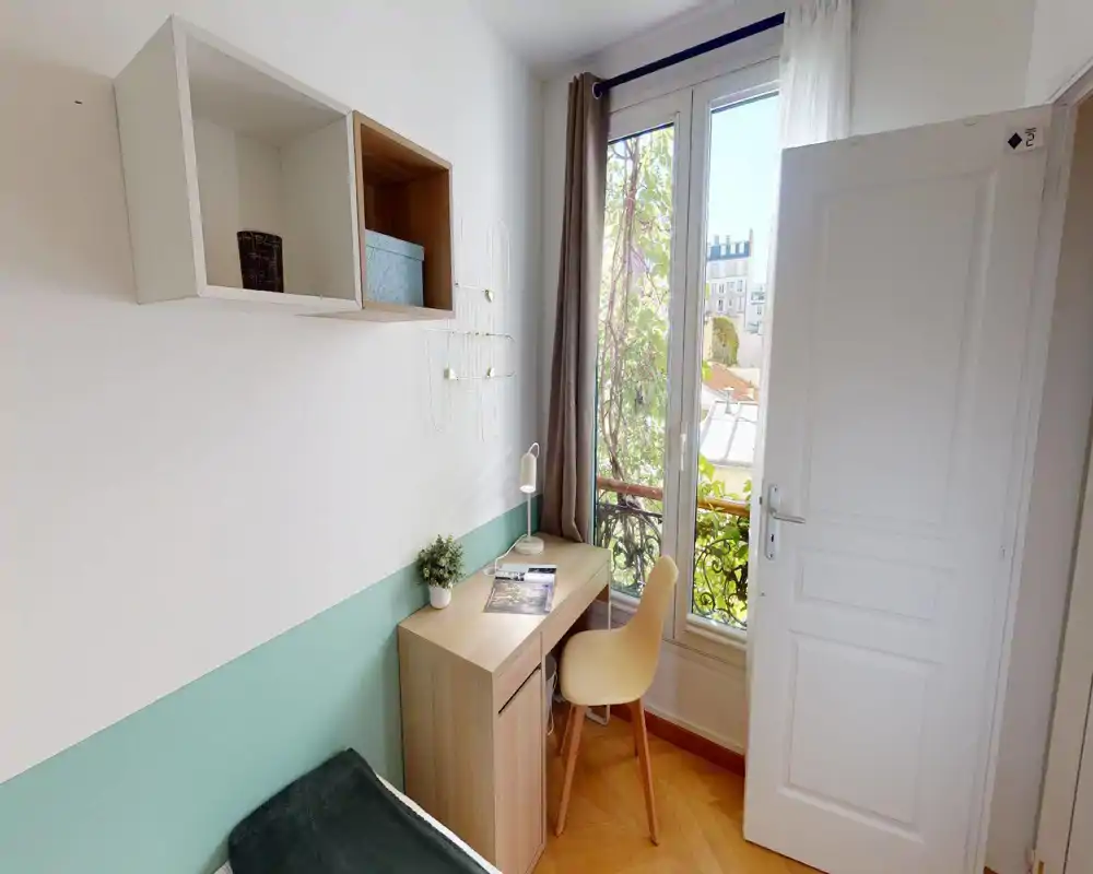Esquina de dormitorio pequeña y luminosa con cama individual, escritorio de madera y una silla junto a la ventana que deja entrar luz natural. Pintura en tonos pastel, estanterías sencillas y plantas decorativas crean un rincón de estudio acogedor.