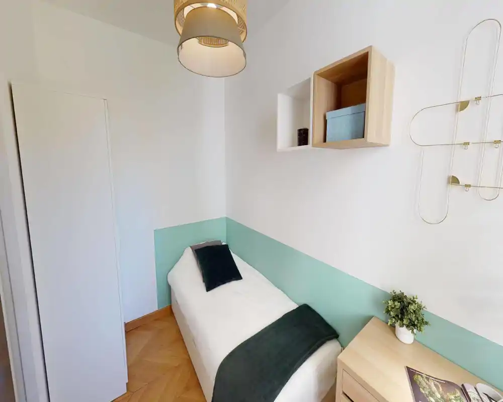 Habitación individual pequeña y bien decorada con cama hecha, franja verde en la pared, muebles de madera clara y buena luz natural.