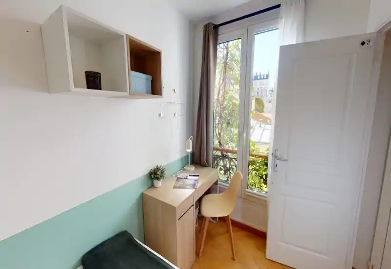 Esquina de dormitorio pequeña y luminosa con cama individual, escritorio de madera y una silla junto a la ventana que deja entrar luz natural. Pintura en tonos pastel, estanterías sencillas y plantas decorativas crean un rincón de estudio acogedor.