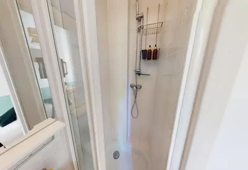 Plato de ducha alicatado y compacto con alcachofa de pared, estante para jabón y puerta plegable de cristal. Espacio de baño brillante, limpio y funcional.