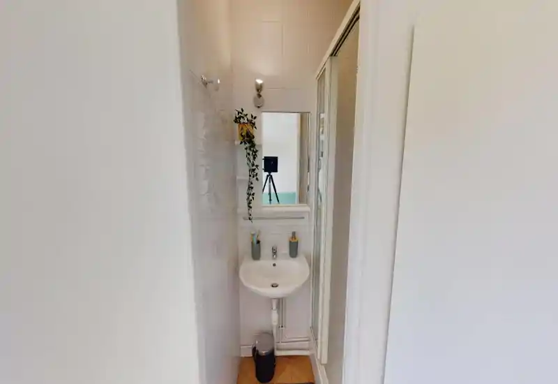 Baño compacto y bien iluminado con un pequeño lavabo, espejo y puerta de ducha. Azulejos blancos limpios, planta colgante y accesorios sencillos que crean un espacio funcional y ordenado.