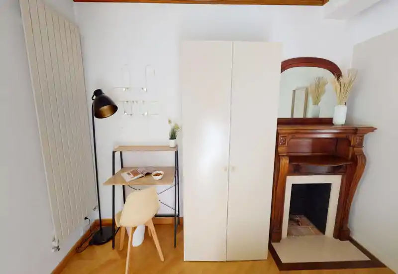 Rincón luminoso y ordenado con un armario, un pequeño escritorio y una silla junto a una chimenea de madera decorativa — ideal para un dormitorio compacto o una zona de estudio.