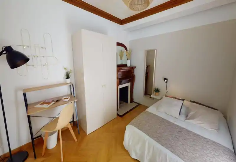 Dormitorio acogedor y luminoso con cama doble, luz de mesilla y pequeño escritorio. Tonos neutros, suelo de madera y chimenea decorativa que aportan calidez y estilo para un anuncio.