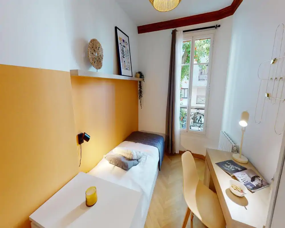 Habitación individual luminosa y bien decorada con pared de acento mostaza, cama cómoda y escritorio compacto junto a la ventana — perfecta para estudiantes o viajeros solos.