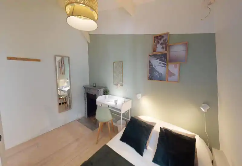 Dormitorio luminoso y cuidado con cama doble, cojines decorativos y un pequeño escritorio junto a una chimenea antigua. Pared verde de acento, cuadros y luz cálida que crean un ambiente tranquilo y acogedor.