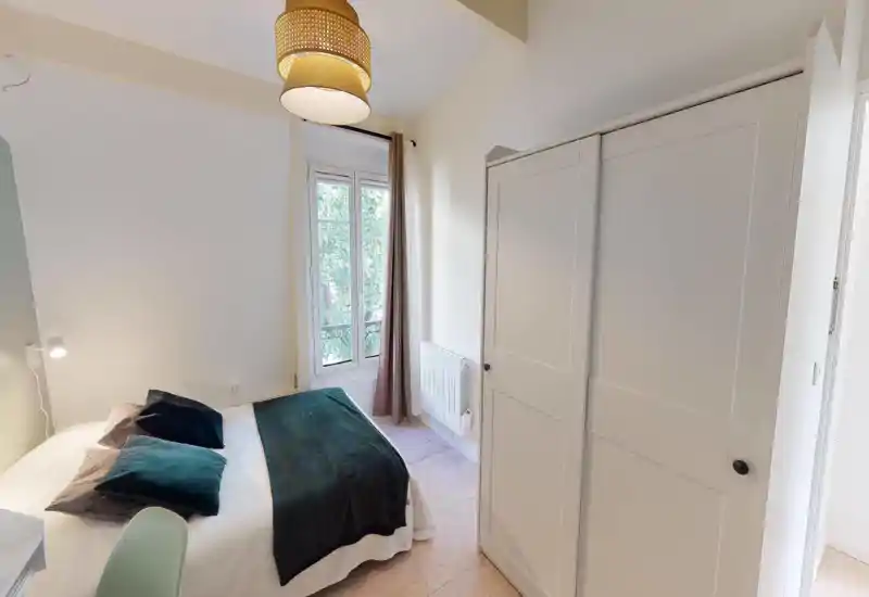 Dormitorio luminoso y acogedor con una cómoda cama doble, cojines suaves y una manta verde oscura. La luz natural entra por una ventana alta con cortinas; un gran armario blanco proporciona amplio almacenamiento.