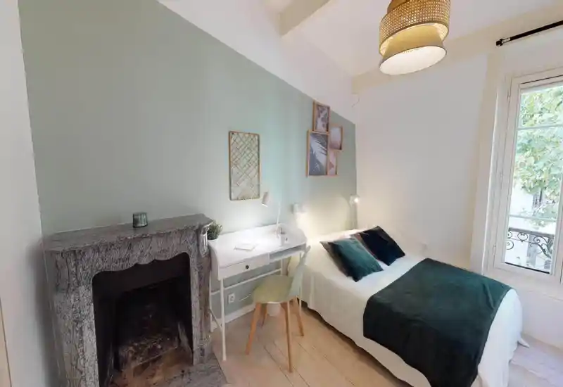 Dormitorio luminoso y bien presentado con cama doble vestida con textiles verde oscuro, lámparas de mesita y un pequeño escritorio blanco junto a una chimenea decorativa y una ventana.