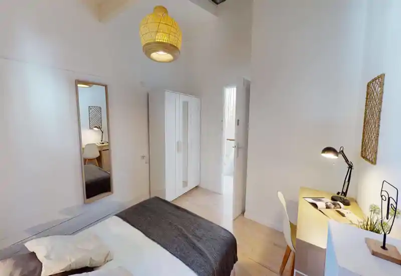 Dormitorio luminoso y minimalista con cama doble, armario y pequeño escritorio. Tonos neutros, suelo de madera y lámpara de techo cálida crean un ambiente tranquilo y acogedor, perfecto para estancias cortas.