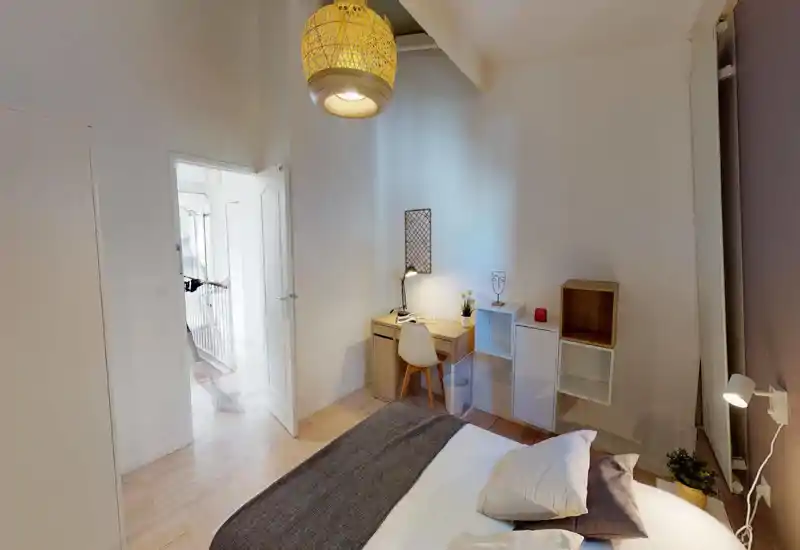 Dormitorio luminoso y acogedor con una cama doble en primer plano, un pequeño escritorio de madera con silla y lámpara en la pared, y una lámpara colgante cálida.