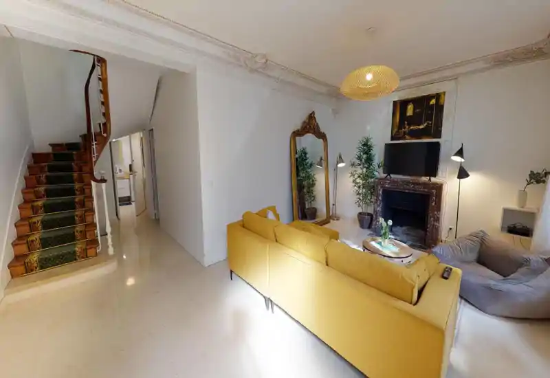Sala de estar espaciosa y con estilo con sofá amarillo mostaza, espejo decorativo, chimenea con televisor, plantas y varias luces — luminosa y acogedora.